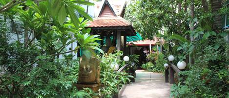 A narrow pathway flanked by lush greenery and a traditional structure with a tiled roof.