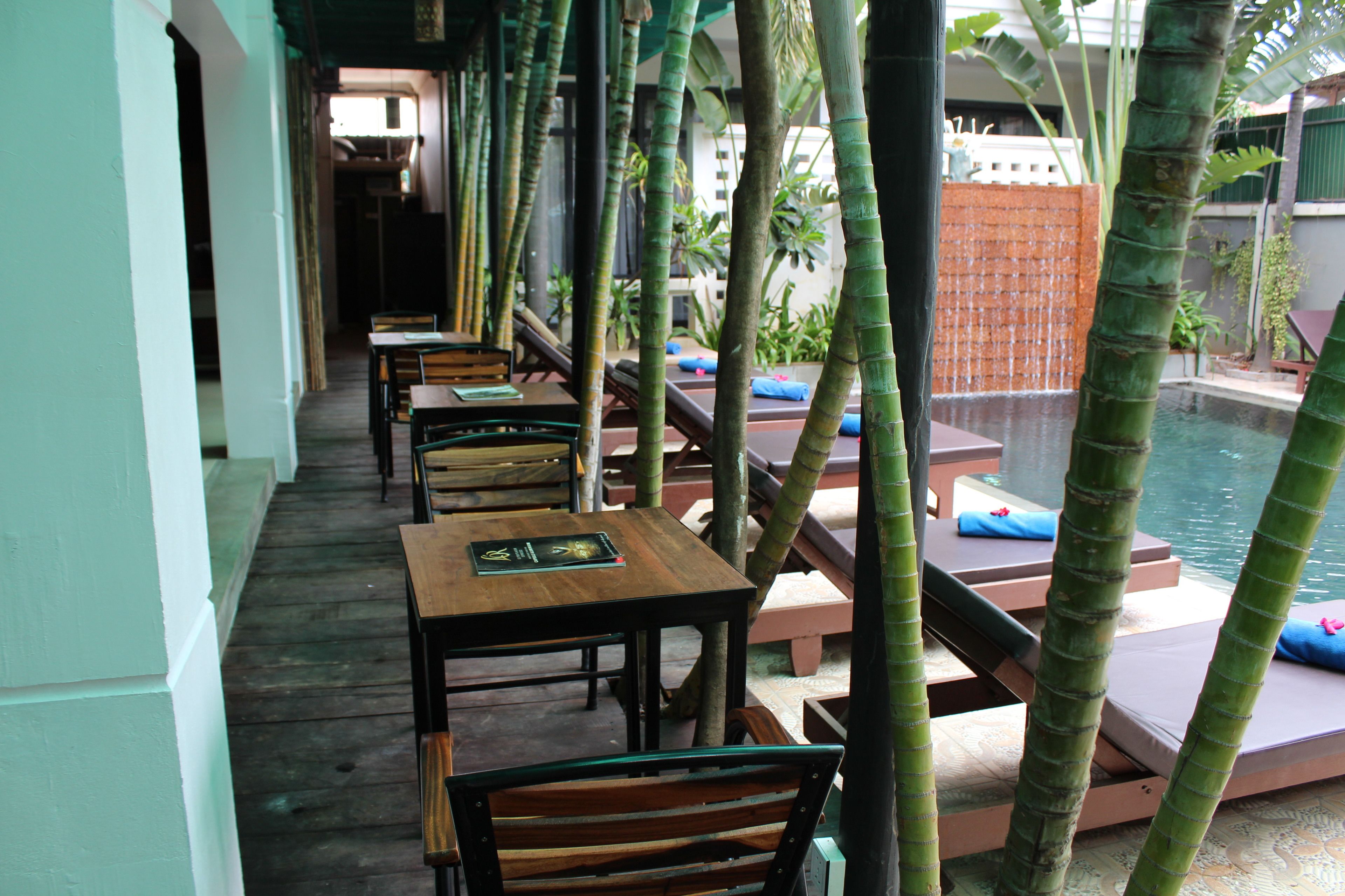 A covered outdoor seating area with wooden tables and chairs, a pool, and palm trees.