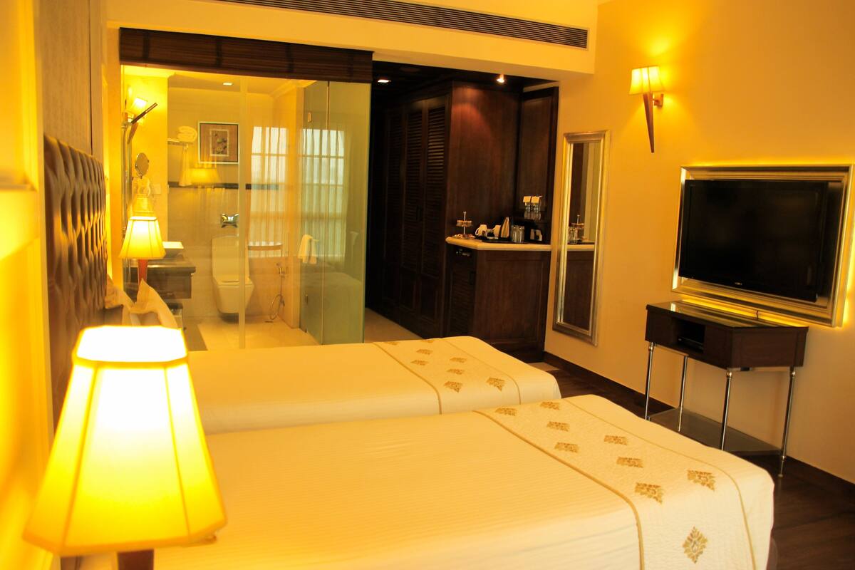 A hotel room with a bed, a TV, a lamp, and a bathroom visible through a glass door.