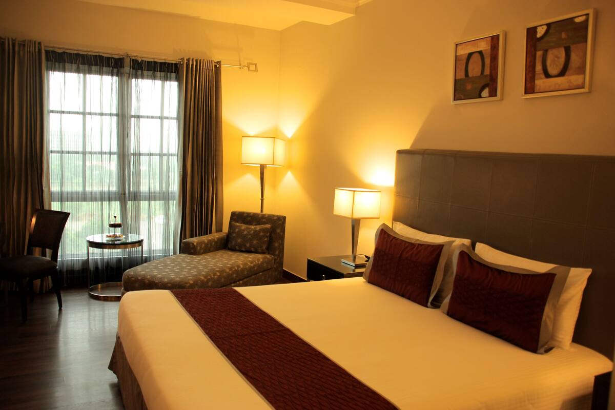 A hotel room with a large bed, two bedside tables, a chair, a small table, and a window with curtains.