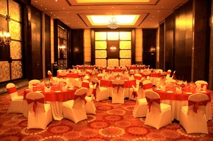 Un salón de banquetes con mesas redondas, sillas blancas adornadas con cintas rojas y una araña de cristal.
