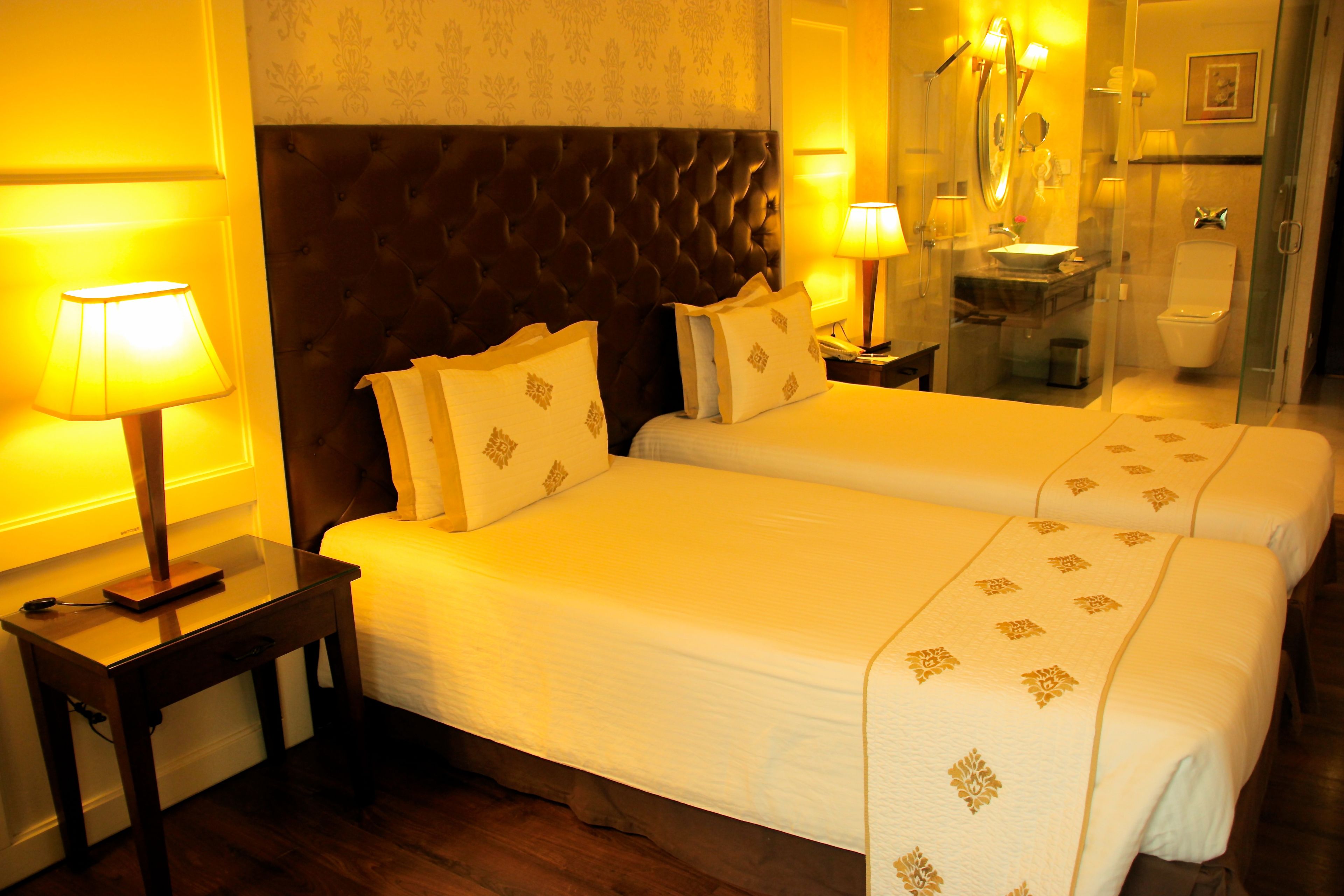 Habitación de hotel con dos camas, cabecero oscuro, lámparas de noche, mesitas de noche y baño con espejo, lavabo e inodoro.