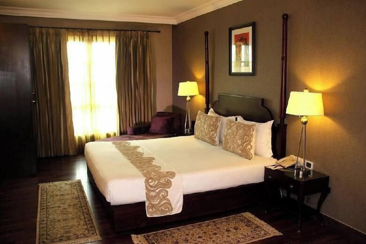 A hotel room with a large bed, two bedside lamps, a nightstand, a bedside telephone, a window with curtains, and a rug.