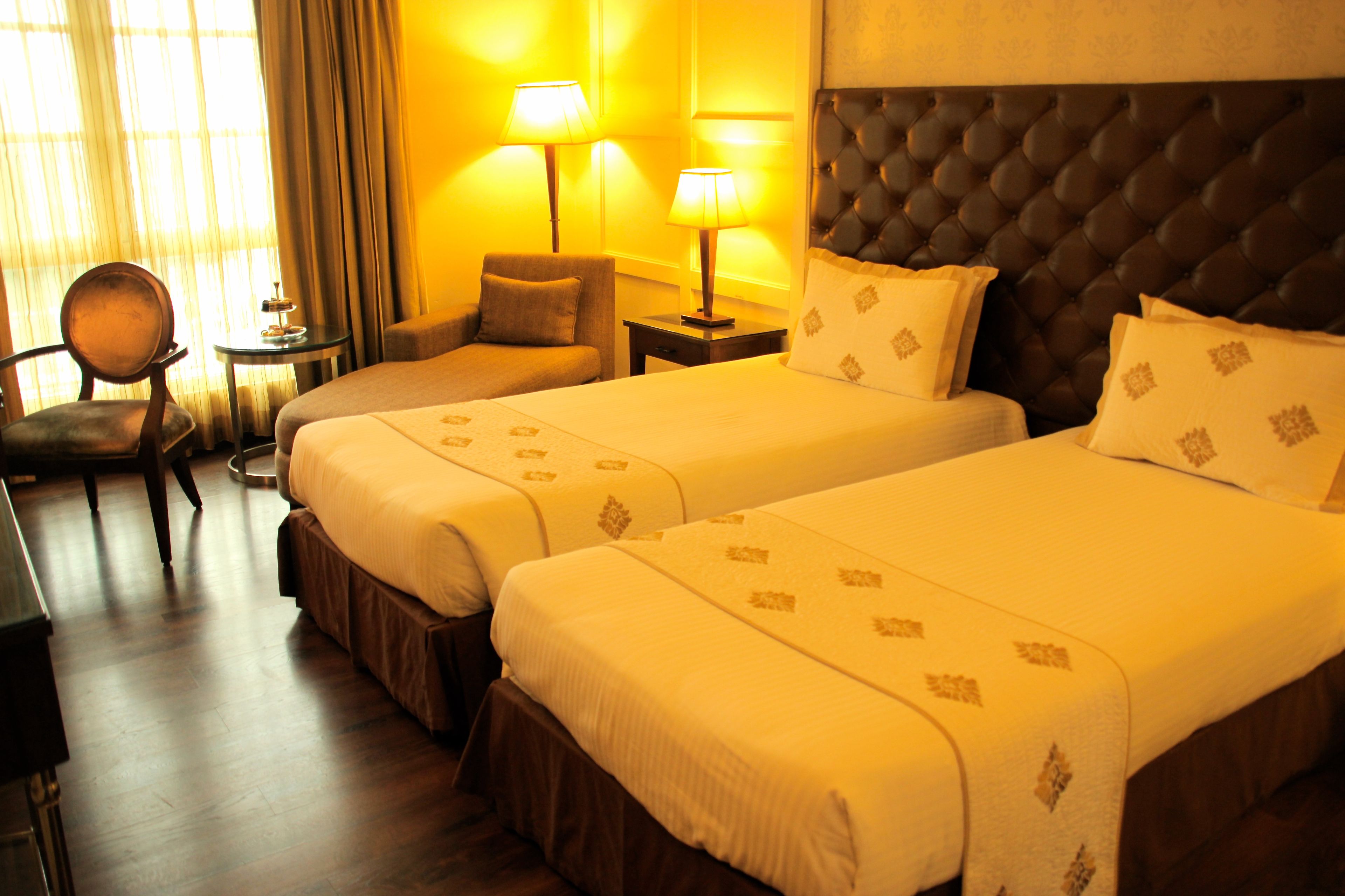 A hotel room with two beds, a chair, a small table, and lamps.