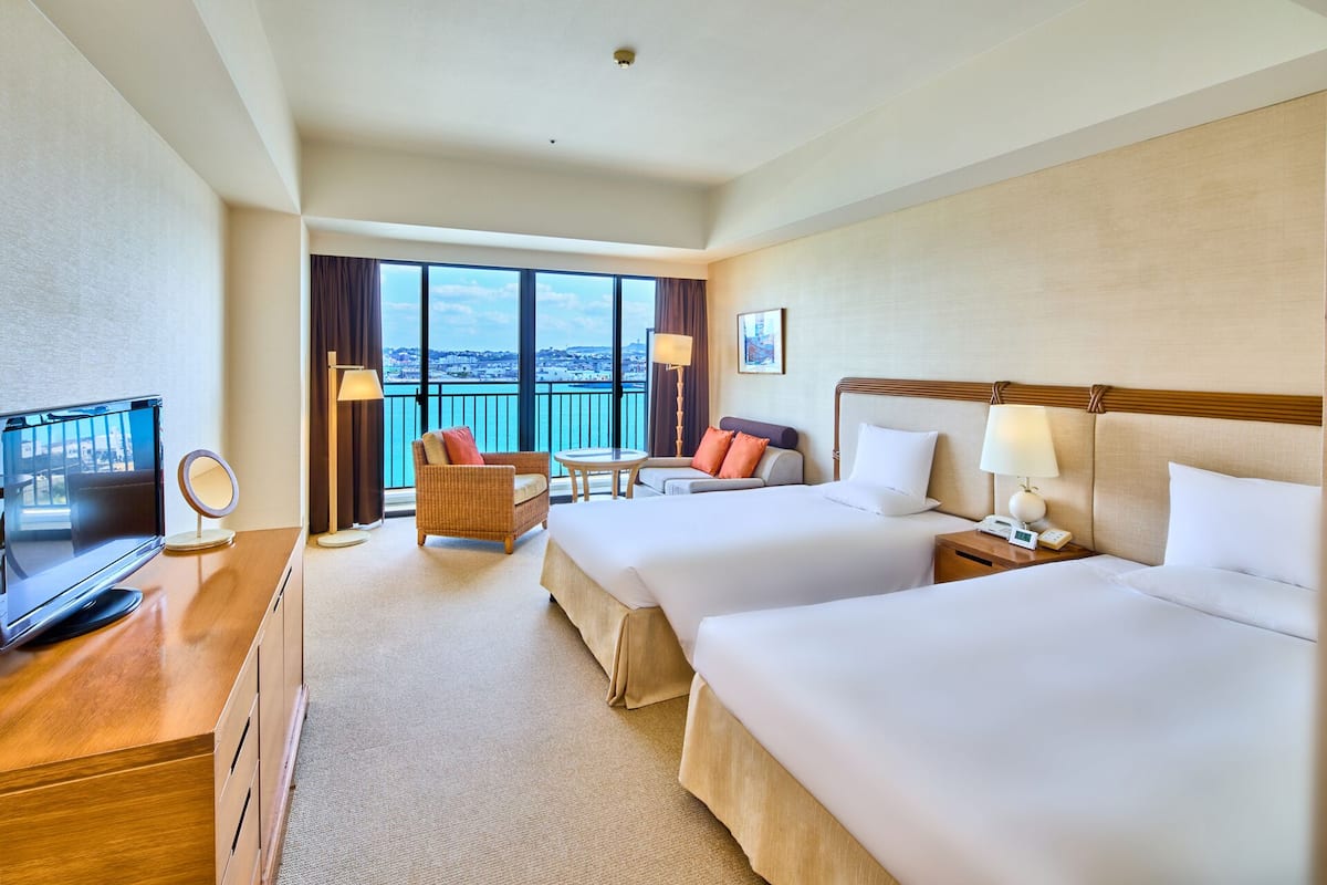A hotel room with two beds, a TV, a desk, a chair, a balcony with a view, and a lamp.