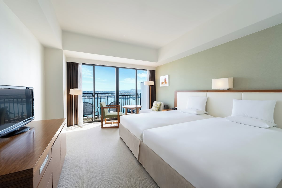A hotel room with two beds, a TV, a desk, a chair, a lamp, and a view of the city.