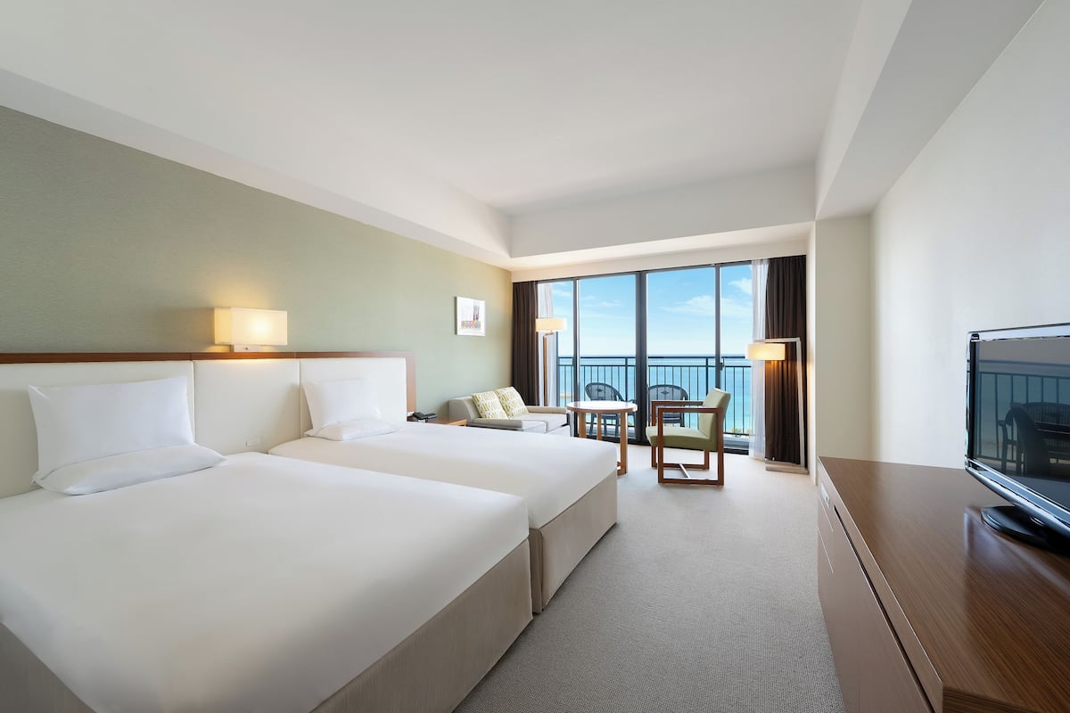 A hotel room with two beds, a TV, a balcony with a view, and a small table with chairs.