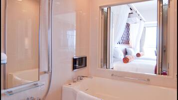 Un baño moderno con ducha acristalada, una bañera grande y un espejo que refleja un dormitorio con cama y cortinas.