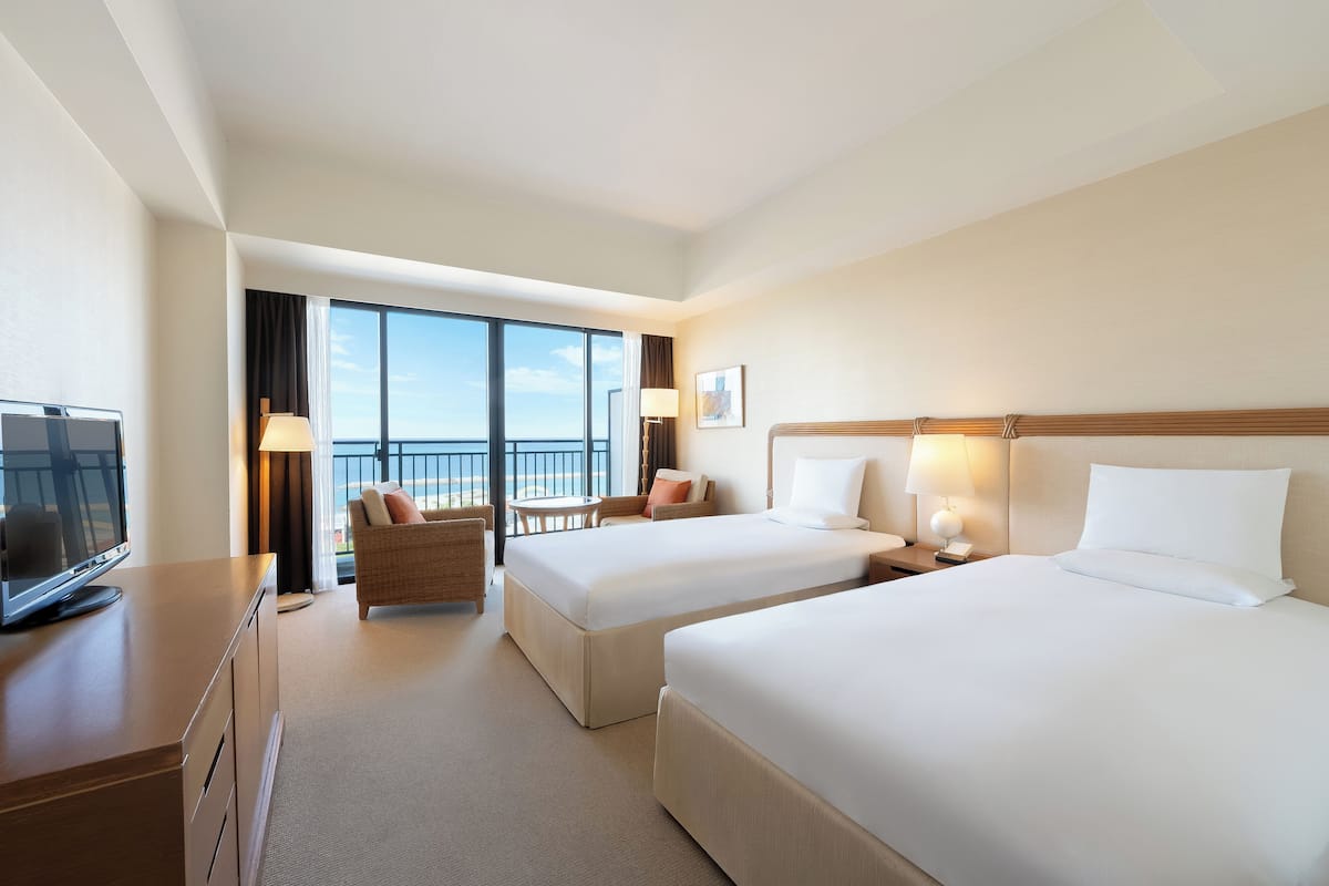 A hotel room with two beds, a desk with a TV, a chair, a lamp, and a view of the sky.