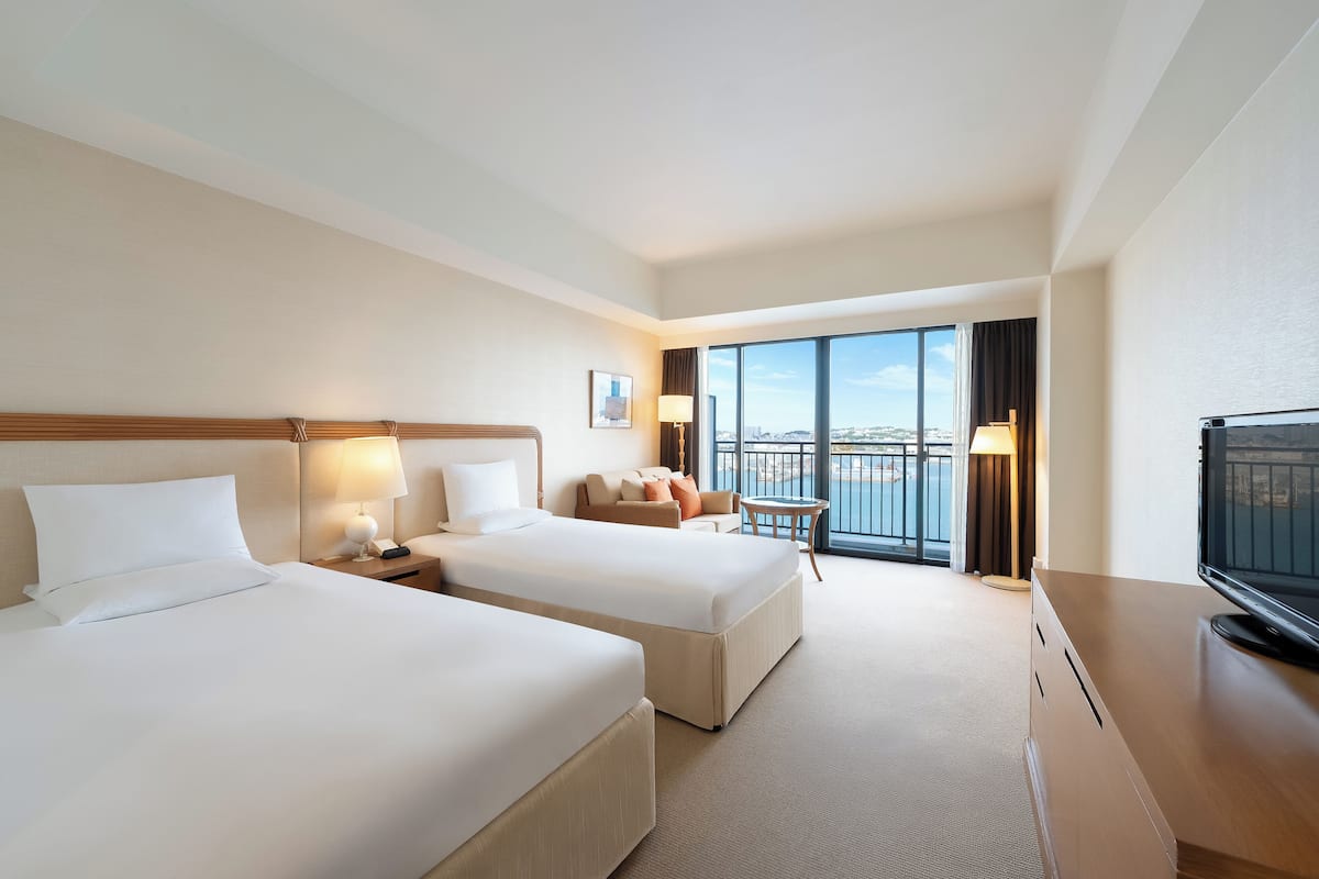 A hotel room with two beds, a TV, a balcony with a view, and a desk with a lamp.