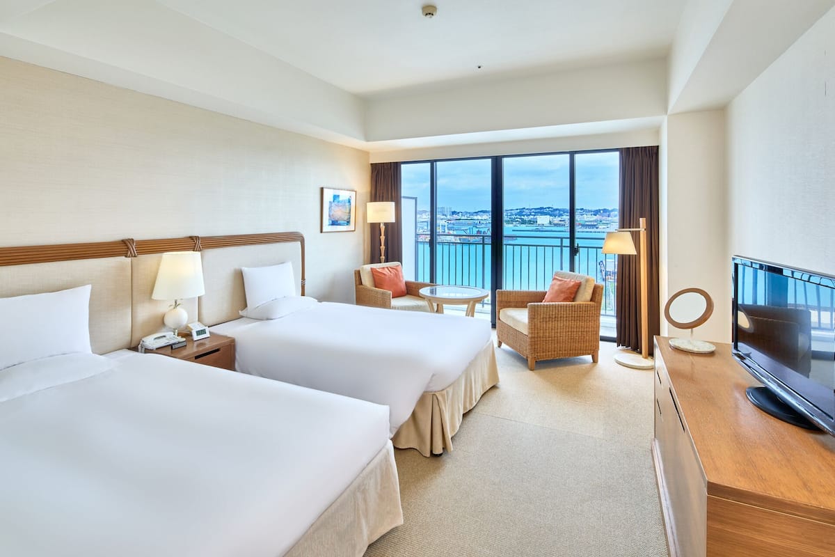 A hotel room with two beds, a balcony with a view, a desk with a TV, and a chair.