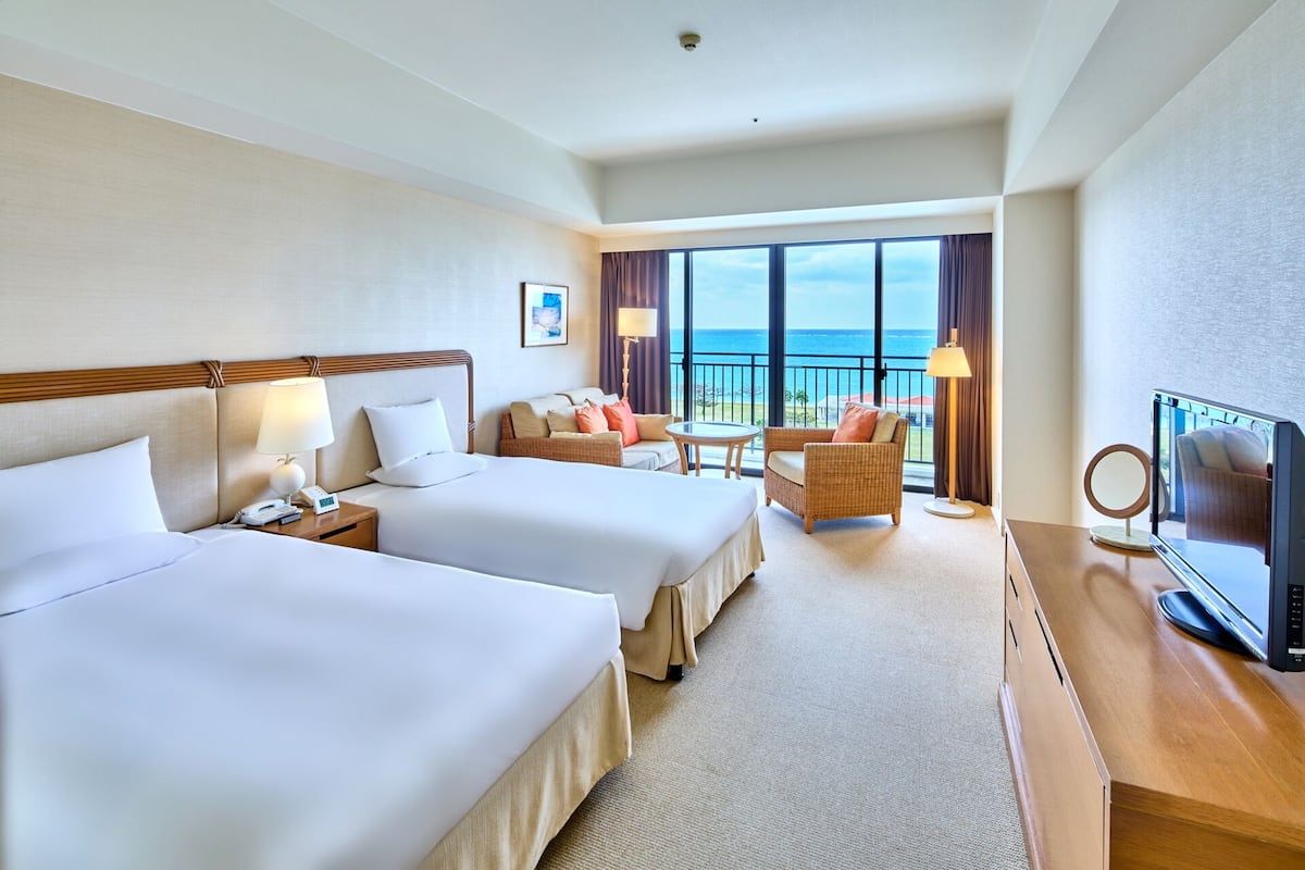 A hotel room with two beds, a sofa, a TV, and a balcony with a view of the ocean.