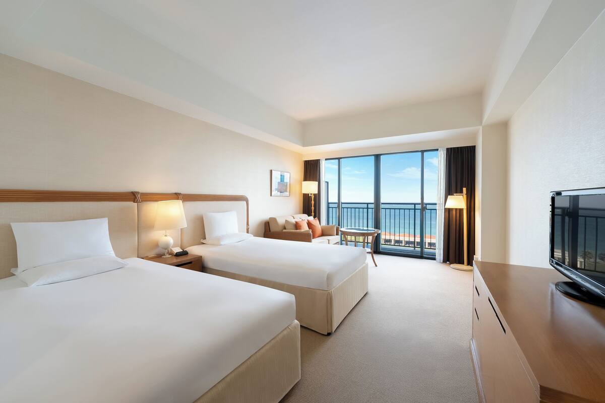 A hotel room with two beds, a television, a balcony with a view, and a desk with a lamp.