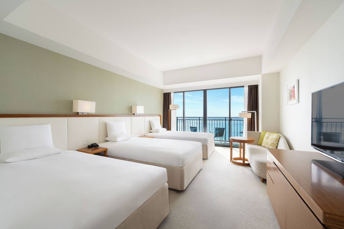 A hotel room with two beds, a desk, a TV, and a balcony with a view.