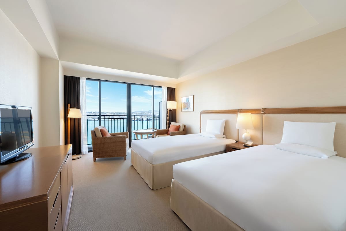 A hotel room with two beds, a TV, a desk, a chair, and a balcony with a view.