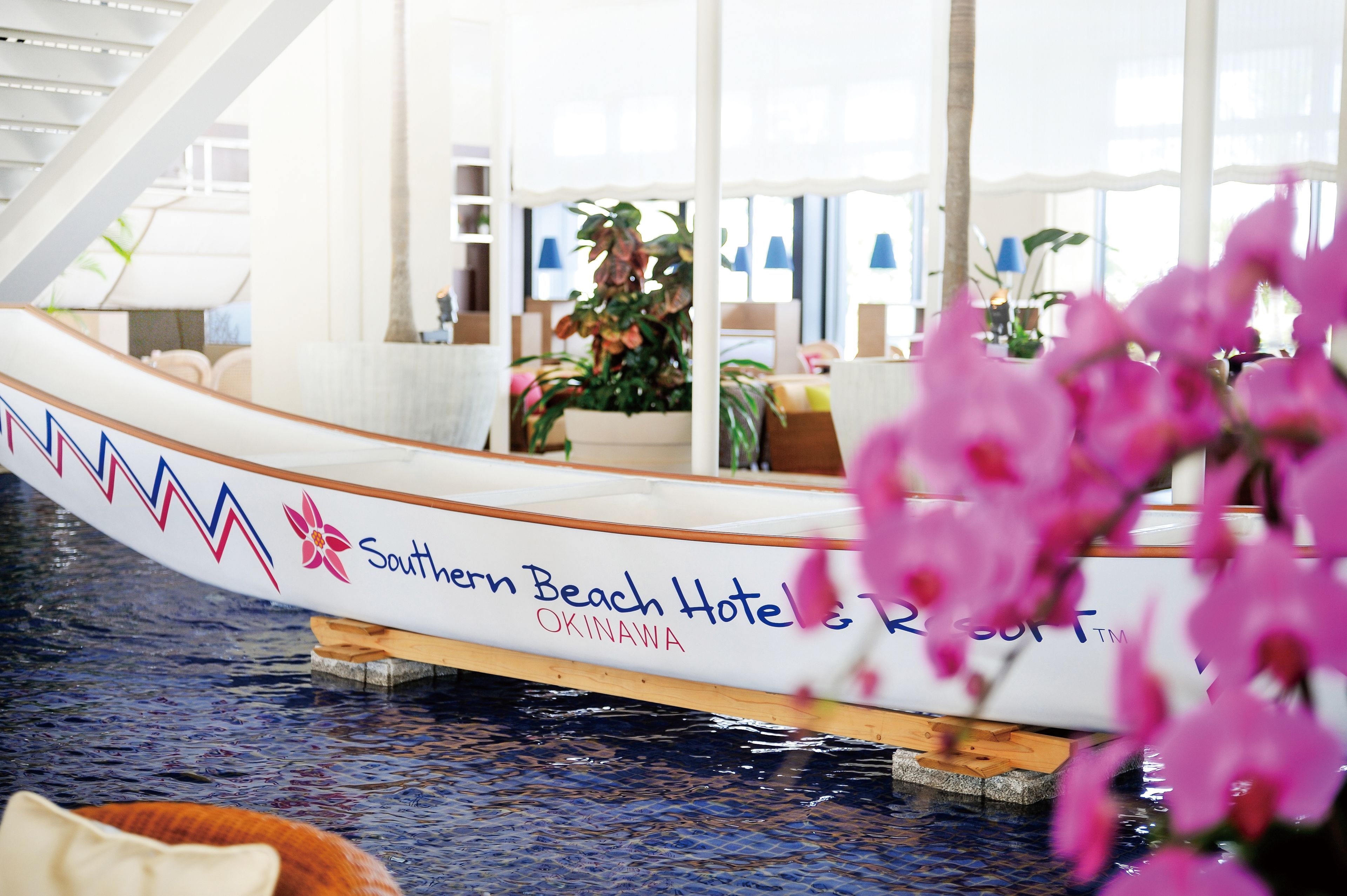 A boat with "Southern Beach Hotel & Resort Okinawa" written on it, docked indoors with a view of a room with a large window and plants.