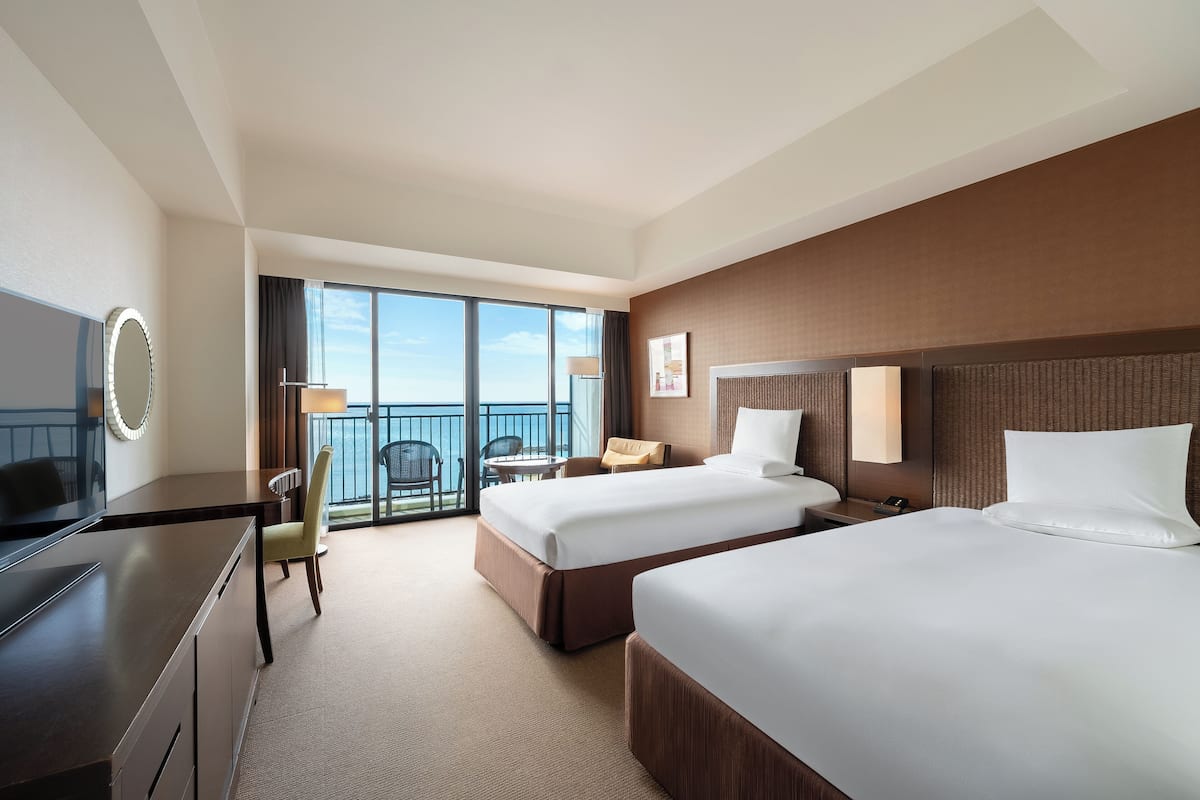 A hotel room with two beds, a desk, a chair, a small table, and a balcony with a view.