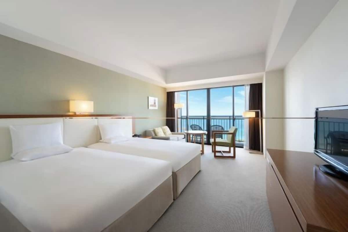 A hotel room with two beds, a desk, a TV, and a balcony with a view.