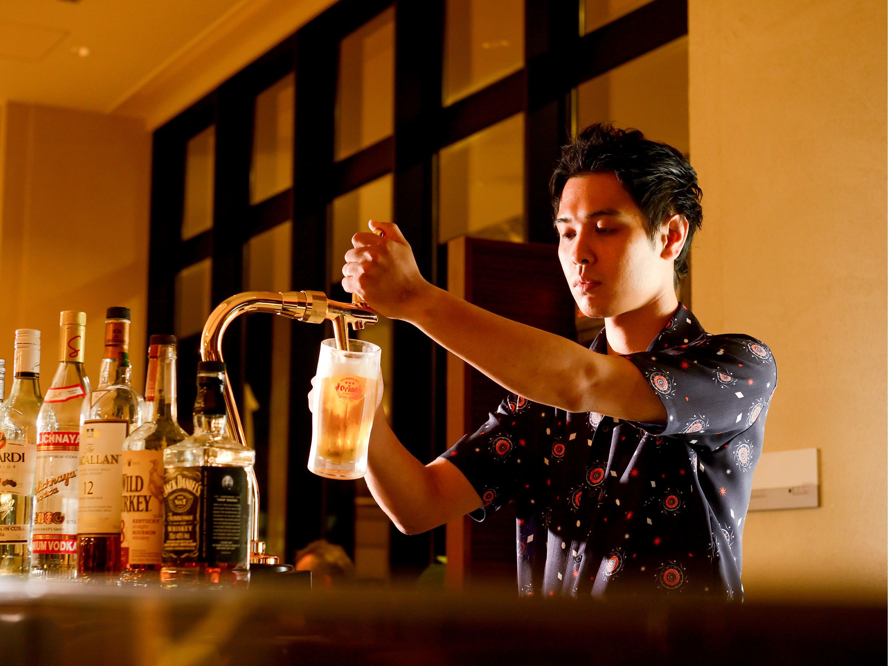 Un barman verse une boisson dans un verre au comptoir d’un bar, où l’on peut voir différentes bouteilles d’alcool.