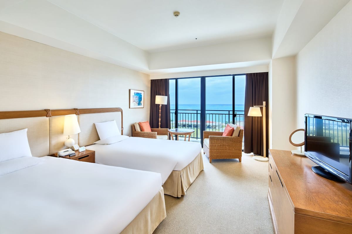 A hotel room with two beds, a TV, a balcony with a view of the sea, and a large window.