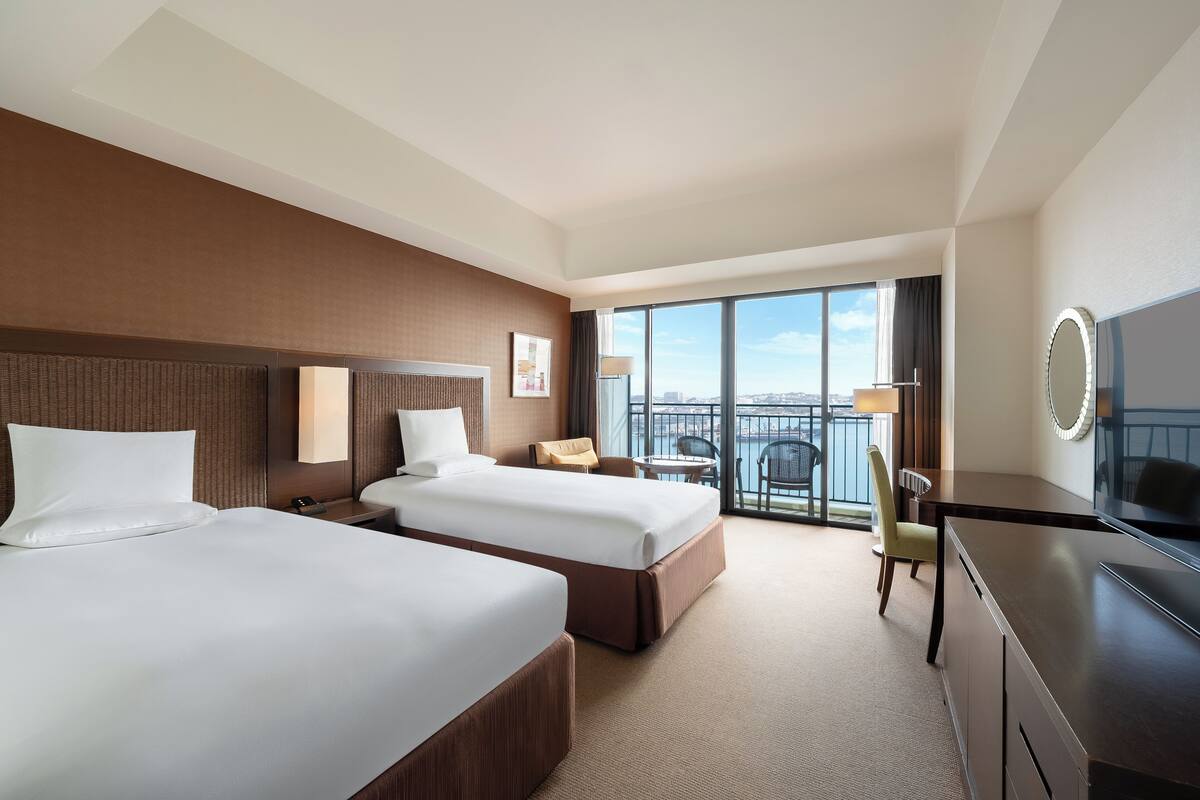 A hotel room with two beds, a desk, a chair, a TV, and a balcony with a view.
