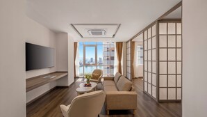 Una sala de estar moderna con un sofá, sillas, un televisor y vistas a un paisaje urbano a través de amplios ventanales.