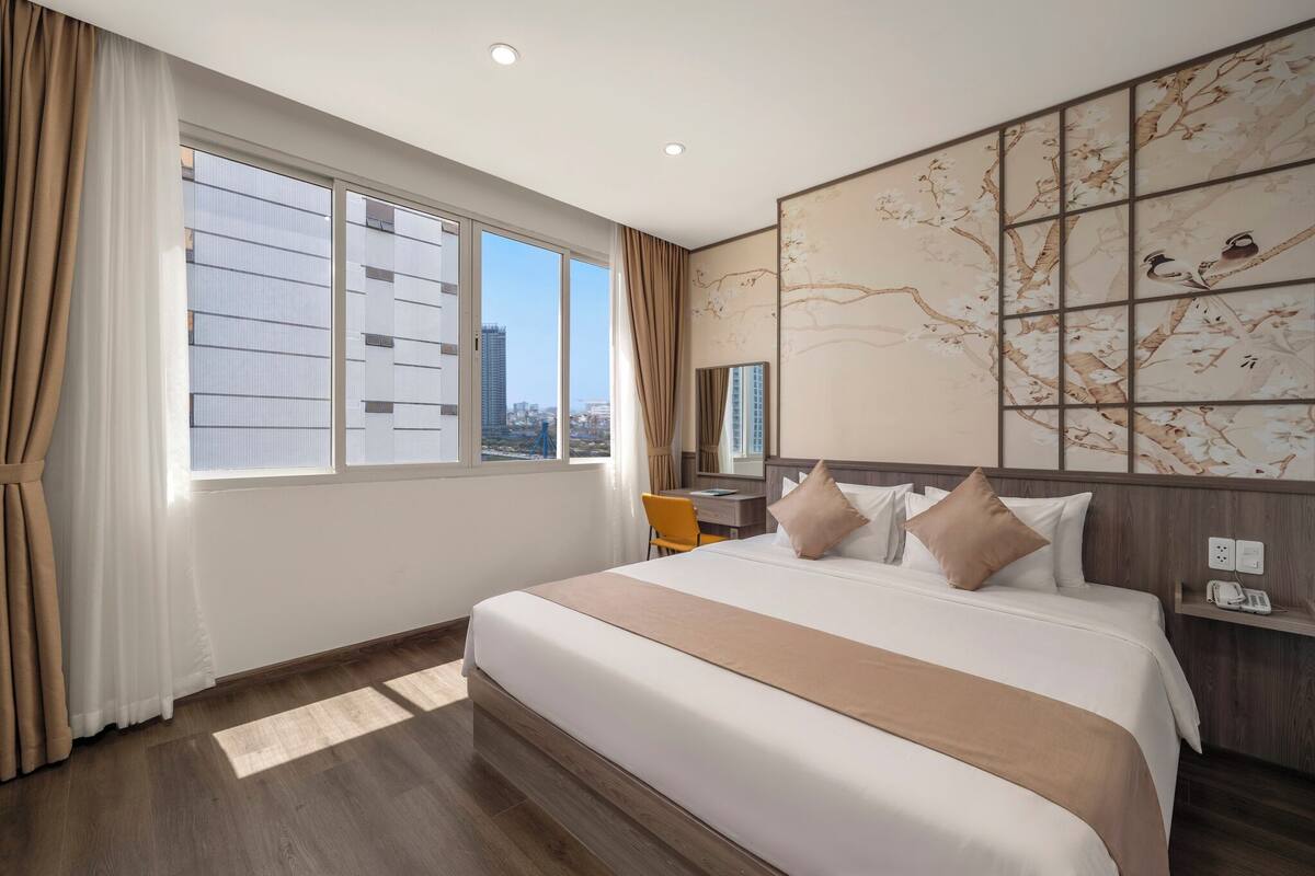 Quarto de hotel com uma cama grande, uma escrivaninha, uma cadeira, uma janela com cortinas e um mural de árvore na parede.