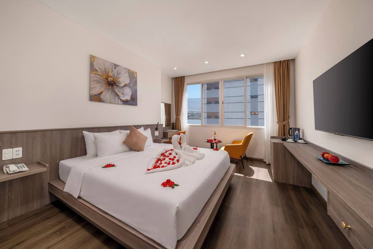 Quarto de hotel com cama, escrivaninha, cadeira, televisão e janela com cortinas.