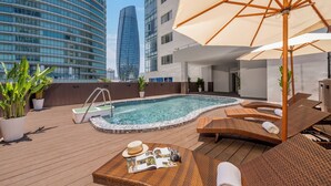 Un área de piscina en la azotea con sillones de descanso, un jacuzzi y un gran paraguas.