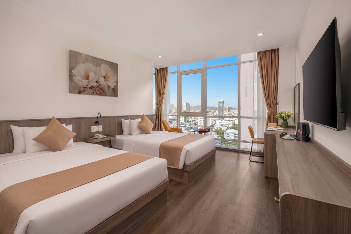 Quarto de hotel com duas camas, televisão, escrivaninha e uma grande janela com vista para a cidade.
