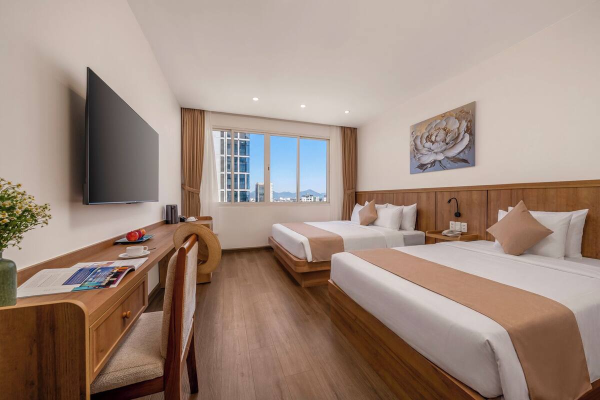 Quarto de hotel com escrivaninha, duas camas, televisão, janela com vista para a cidade e um quadro na parede.
