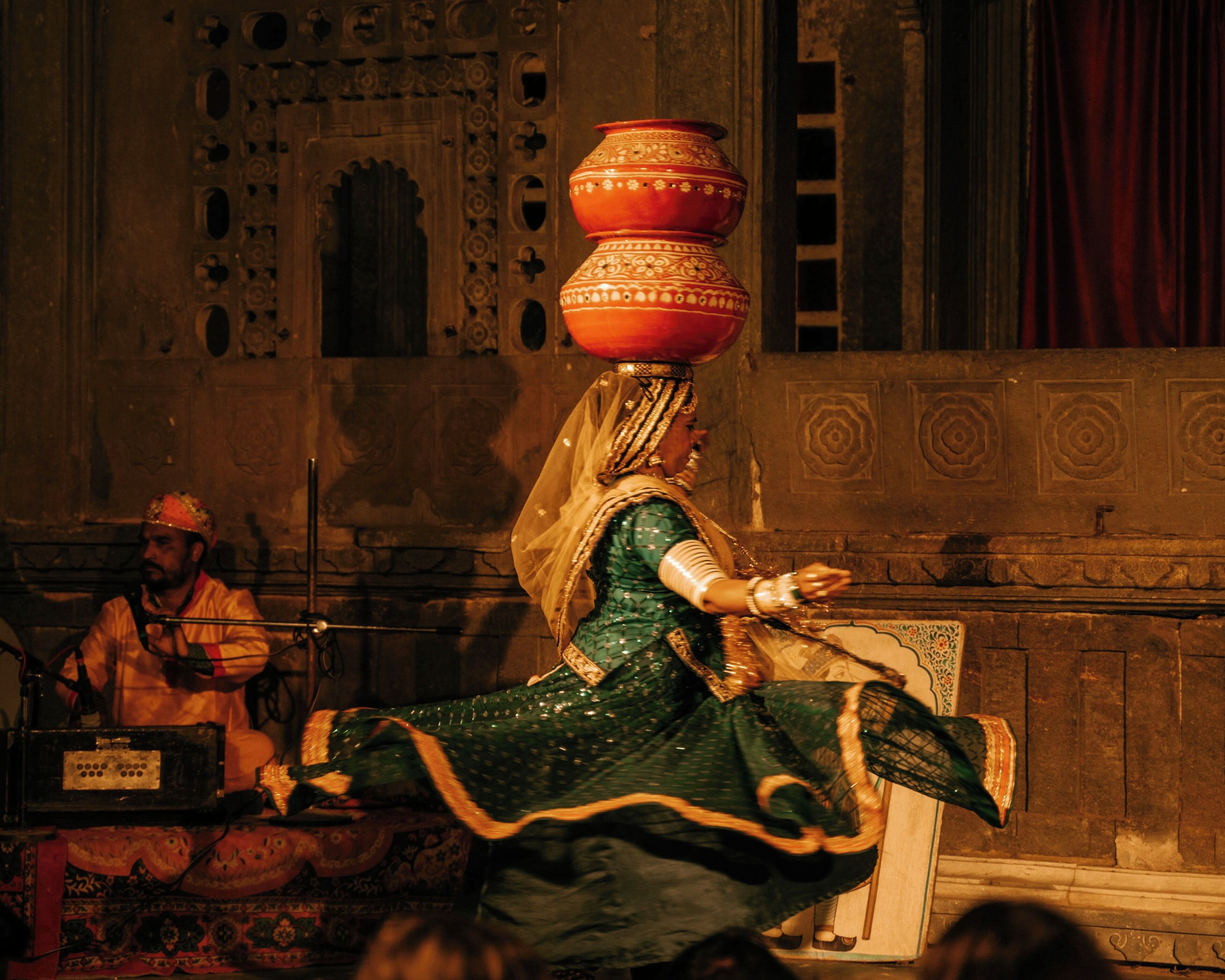 Una presentación de danza tradicional con una bailarina vestida de verde y una olla roja en la cabeza, frente a una arquitectura ornamentada.