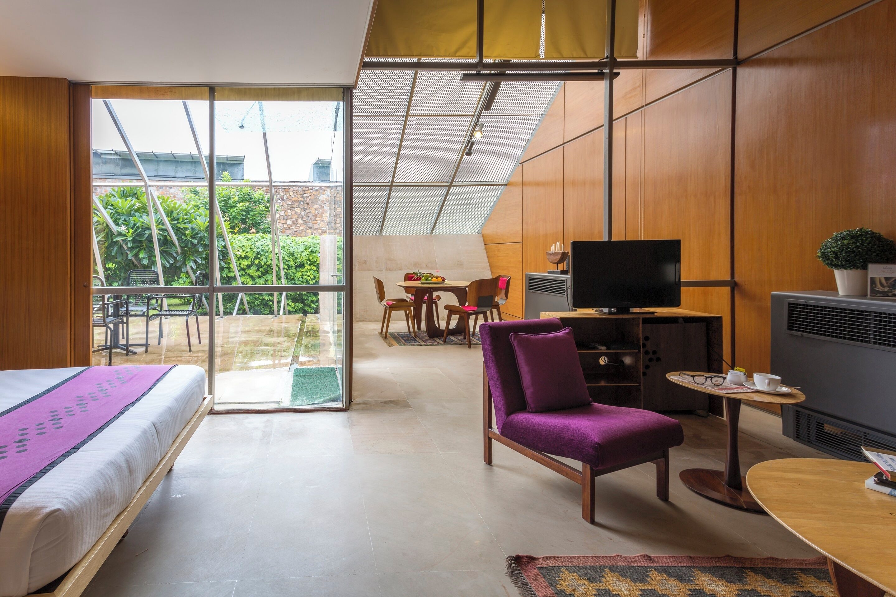 Una habitación de hotel moderna con una cama, un sillón morado, una mesita, un televisor y un balcón con vistas a la vegetación.