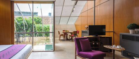 Una habitación de hotel moderna con una cama, un sillón morado, una mesita, un televisor y un balcón con vistas a la vegetación.