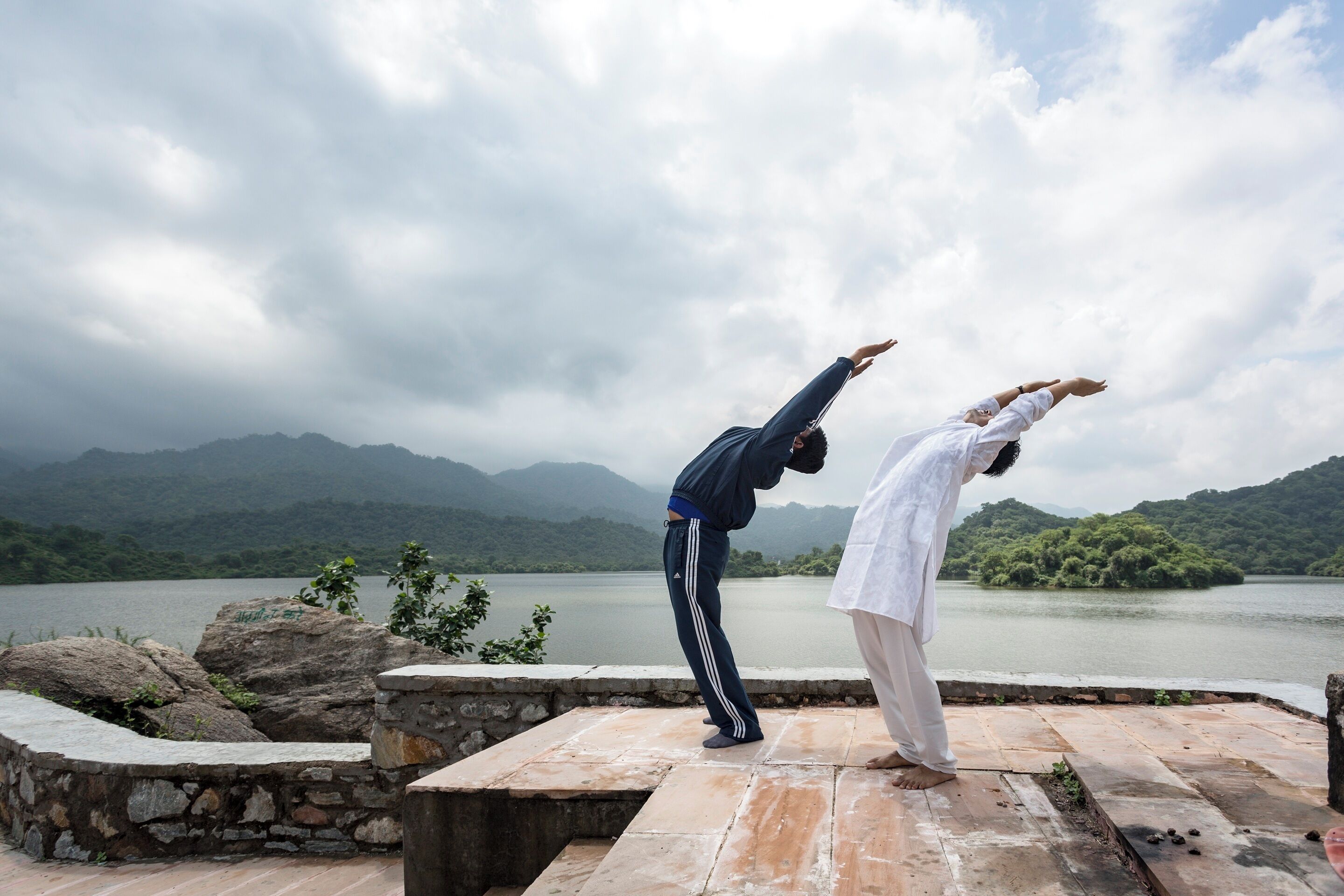 Dos personas practicando yoga sobre una plataforma de piedra, con un lago y montañas al fondo.