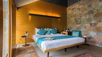 A modern hotel room with a large bed, wooden headboard, stone wall, and a small side table.