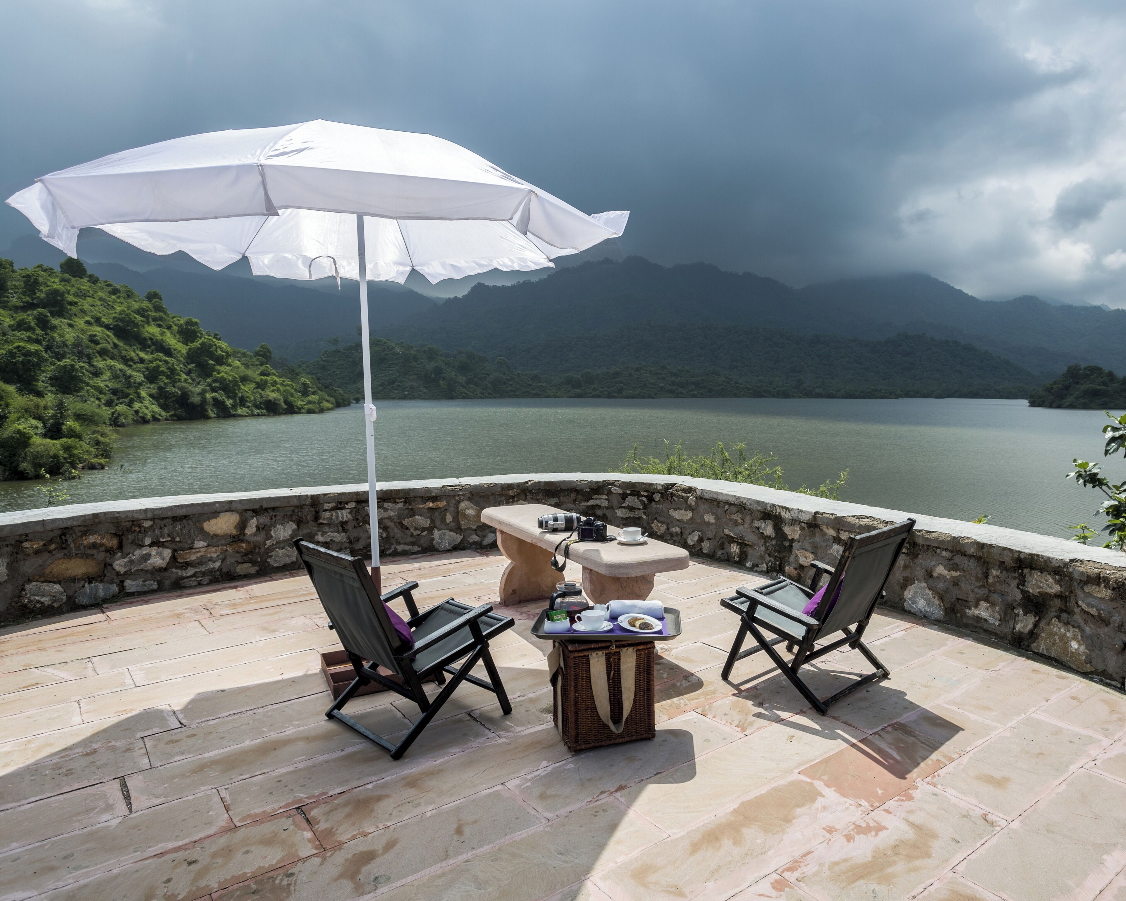 A terrace with two chairs, a table, and a white umbrella overlooking a lake and mountains.