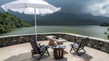 A terrace with two chairs, a table, and a white umbrella overlooking a lake and mountains.