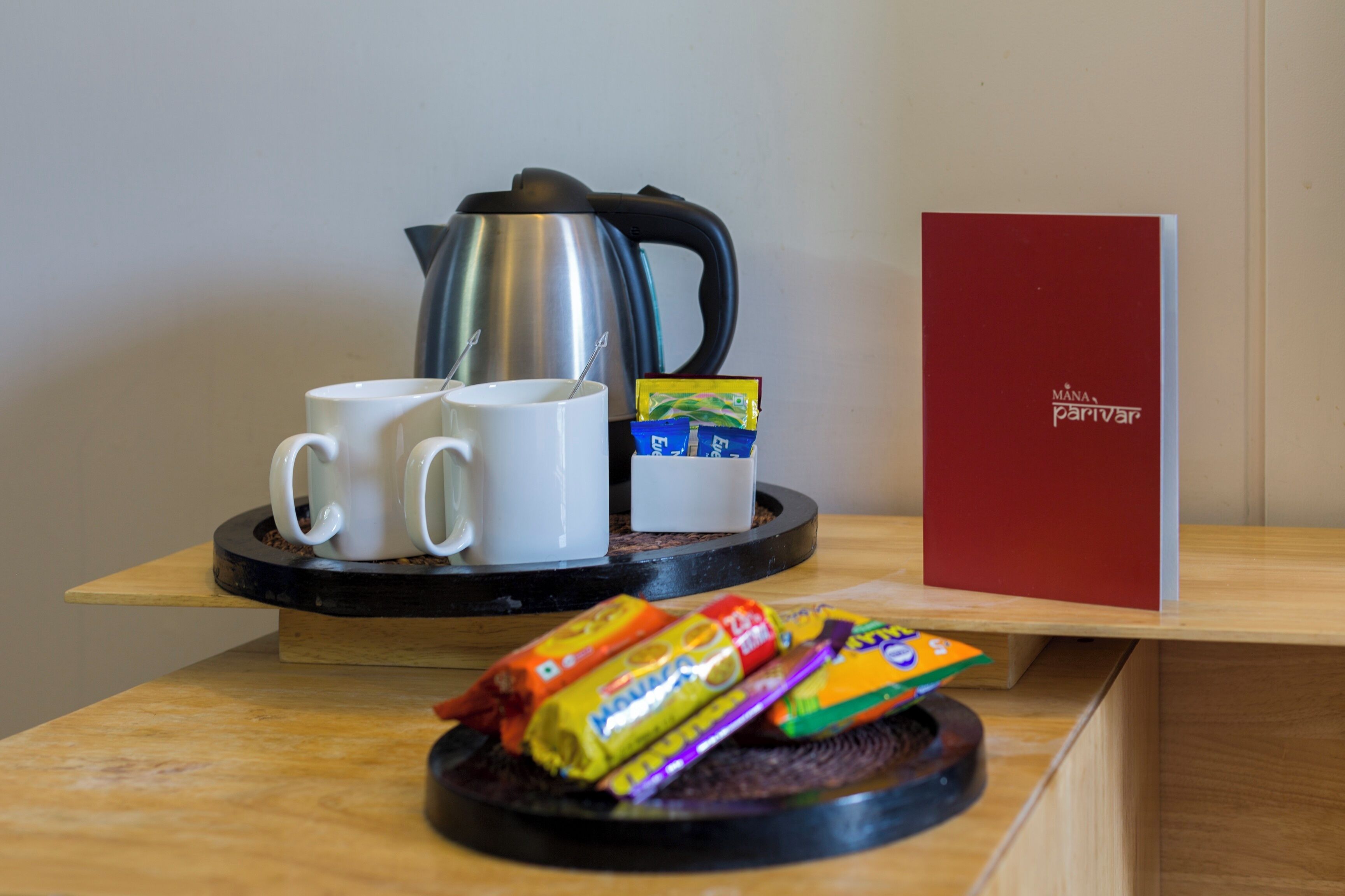 A coffee station with two white mugs, a kettle, and a red book titled 'MANA parivar' on a wooden table.