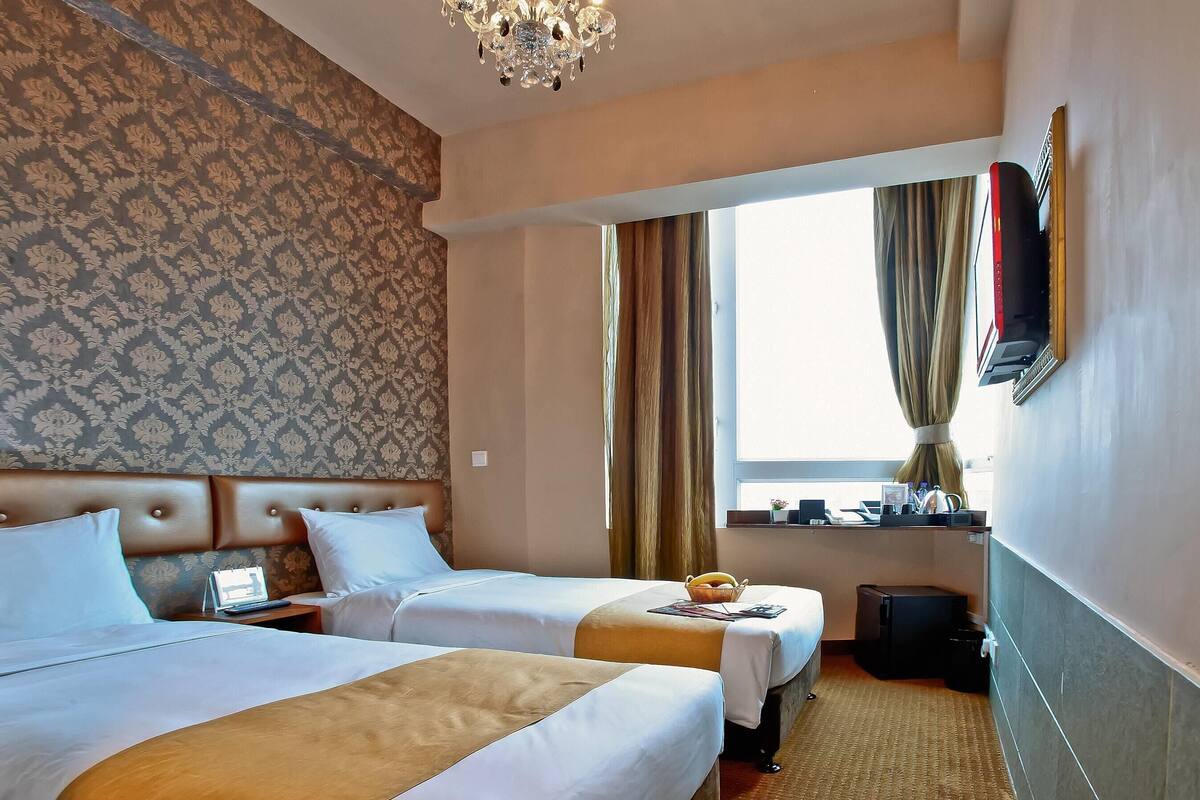 A hotel room with two beds, a chandelier, a window with curtains, a flat-screen TV, and a small table with a basket.