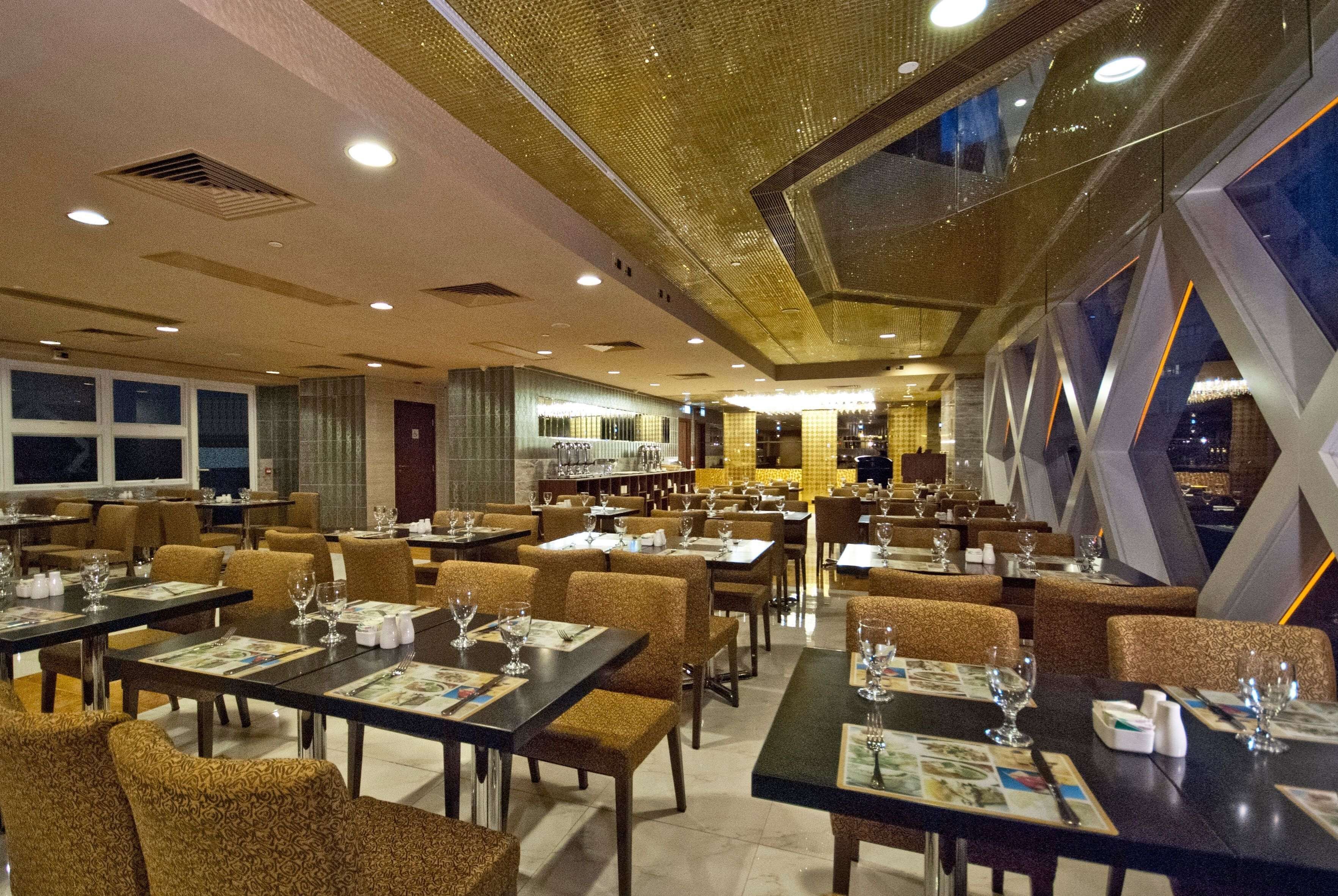 A spacious restaurant with multiple dining tables and chairs, a bar area, and large windows offering a view of the cityscape.