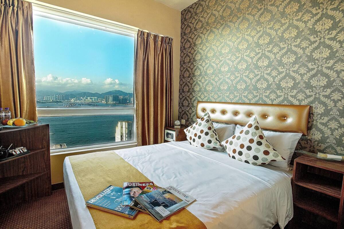 A hotel room with a large window, a bed with a tufted headboard, and a nightstand with a clock.