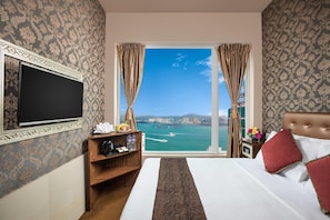 A hotel room with a bed, a TV, a desk with a coffee maker, a shelf with toiletries, and a view of the sea.