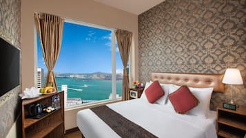 A hotel room with a large bed, a nightstand, a TV, a window with curtains, and a view of the city.