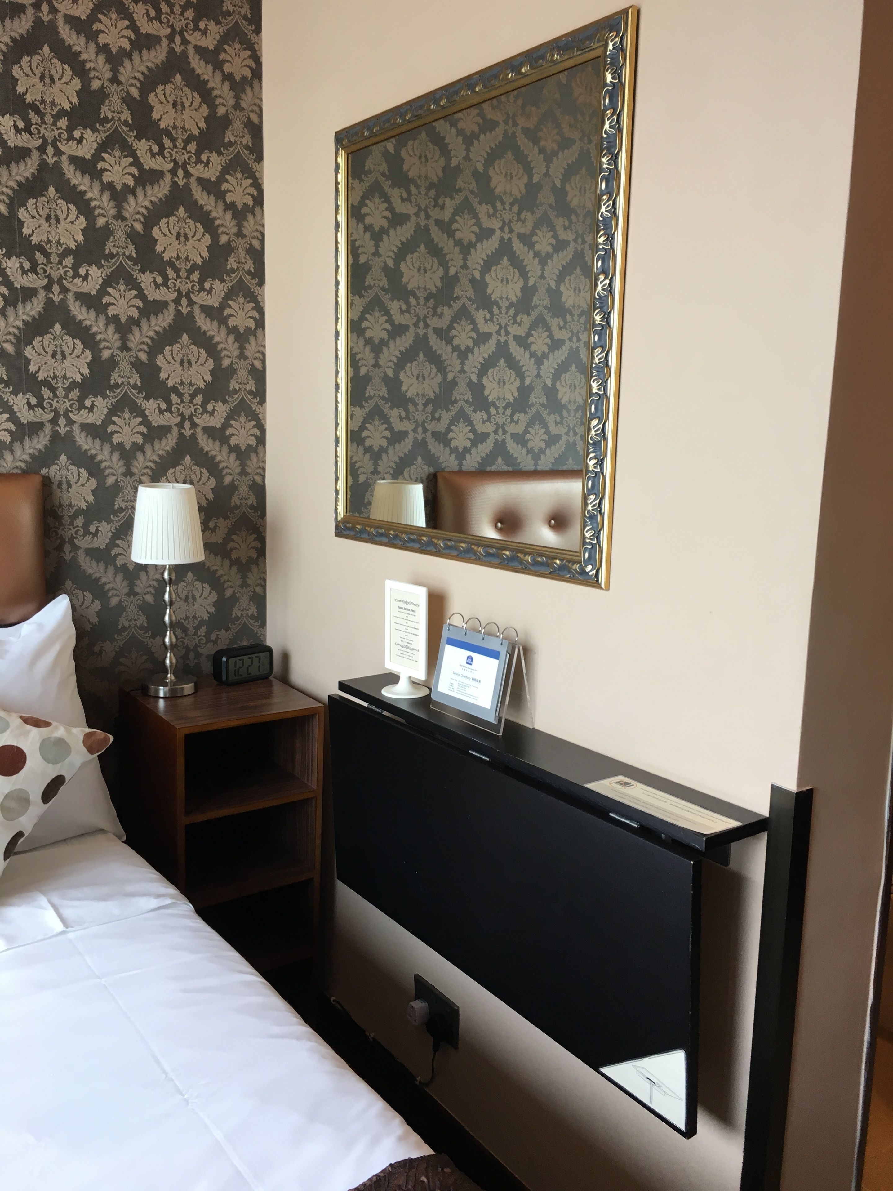 A hotel room with a bed, a nightstand with a lamp, a dresser, a mirror, and a wall clock.