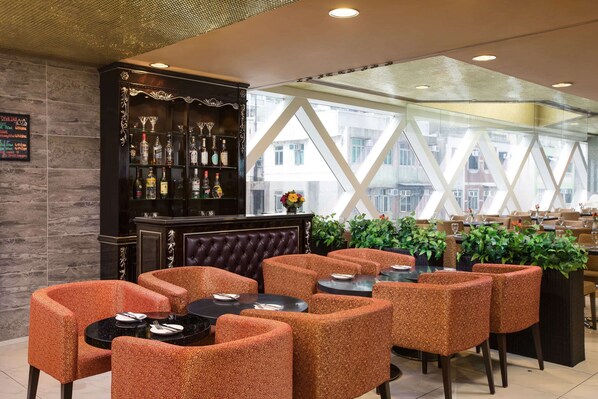Un restaurante con zona de bar que presenta un mueble de madera oscura con estantes de vidrio que exhiben botellas y vasos. Hay varias mesas con sillas de estampado naranja, y el espacio está bien iluminado por luz natural que entra por grandes ventanales.
