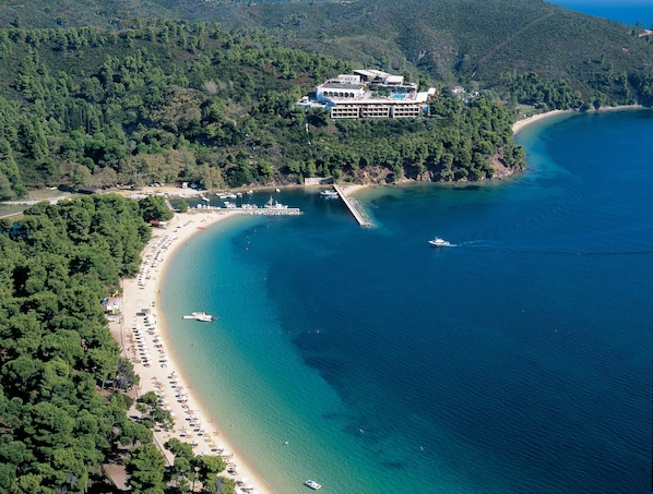An aerial view of a coastal resort with a long sandy beach, a pier, and a large building with multiple balconies overlooking the sea. - Villa Anna Maria (Skiathos)
