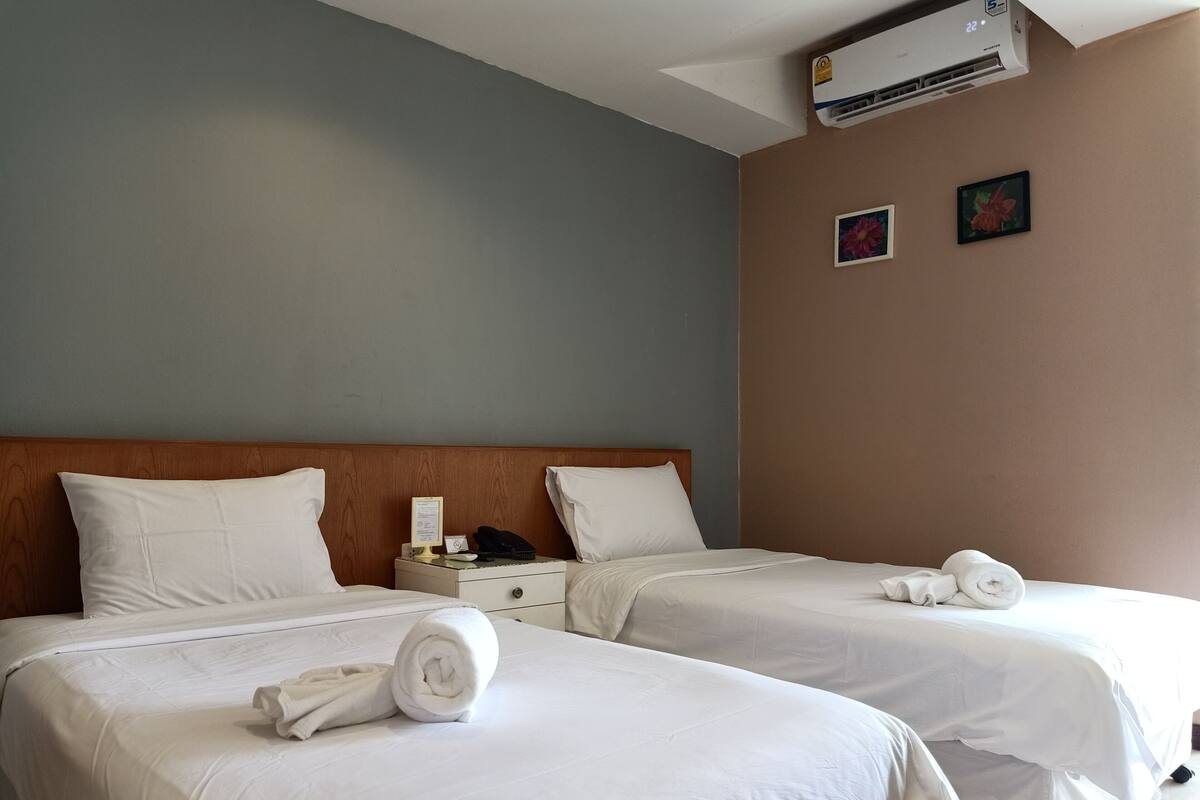 A hotel room with two beds, white bedding, a wooden headboard, a nightstand with a phone and a lamp, and an air conditioning unit on the wall.