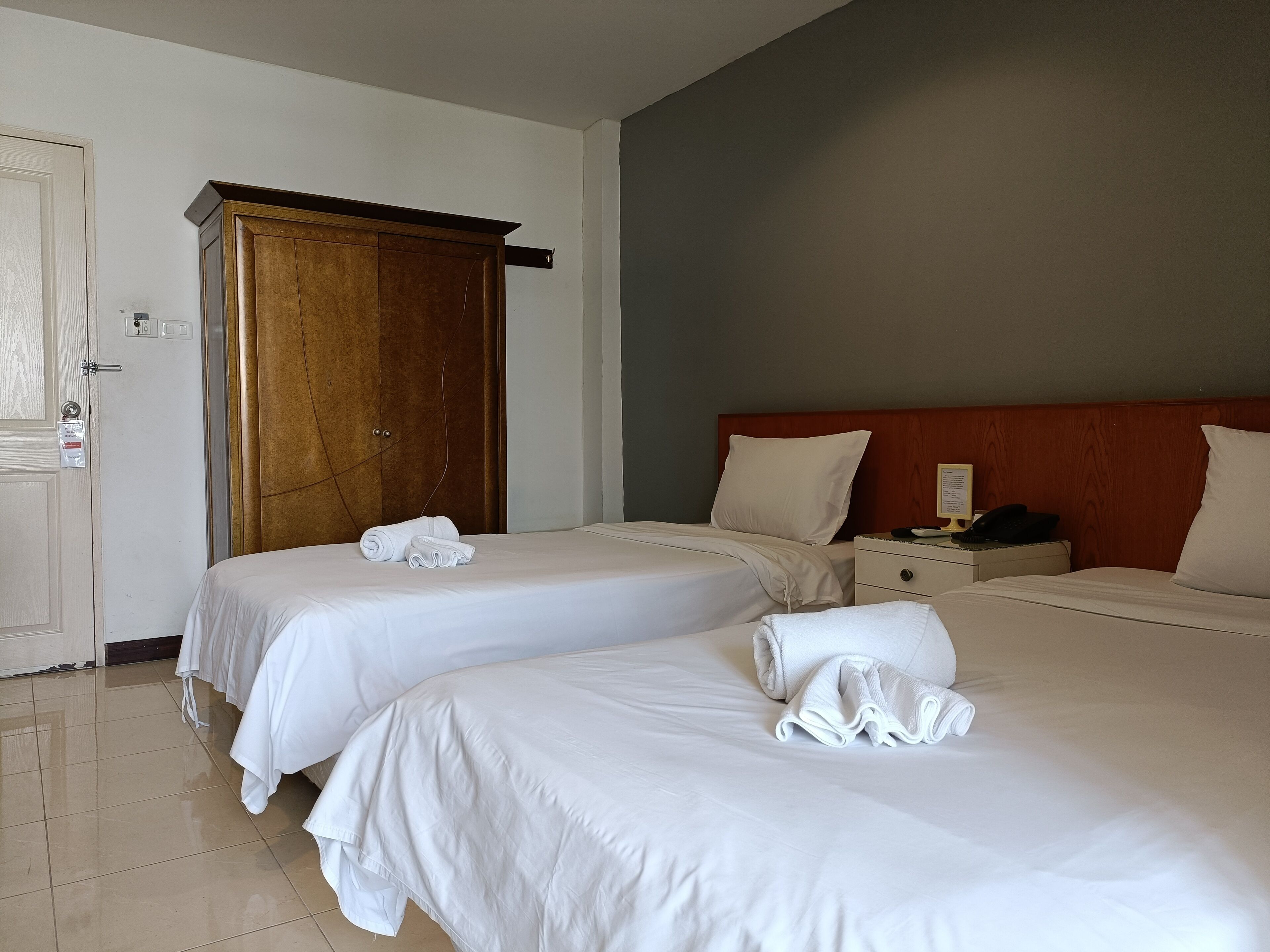 A hotel room with two beds, a wooden wardrobe, a bedside table with a lamp, and a door with a lock and a key card reader.