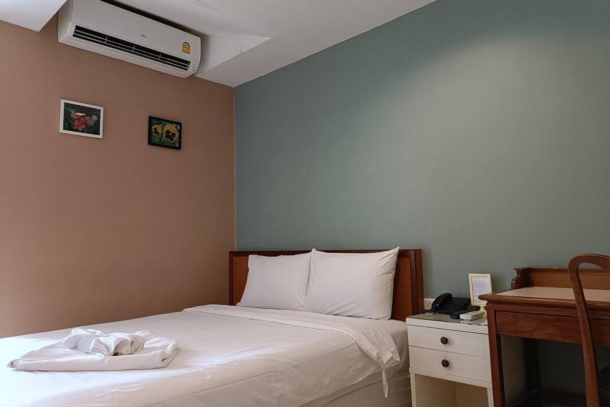 A neatly made bed with white linens, a wooden headboard, a bedside table with a phone and a lamp, and a desk with a chair.